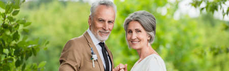 happy middle aged groom in suit holding hands with mature bride in garden, bannerの写真素材