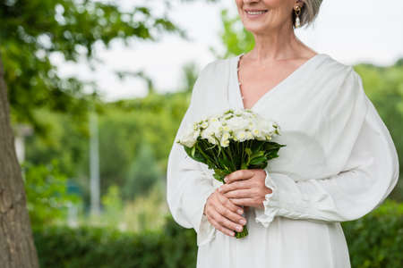 cropped view of happy middle aged bride in white dress holding wedding bouquet in green gardenの写真素材
