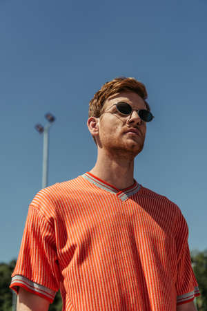 low angle view of redhead man in sunglasses and orange t-shirt under blue sky outdoorsの写真素材