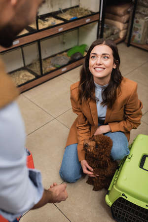 Smiling woman petting poodle near cage and blurred boyfriend in pet shop