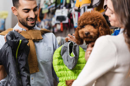 Smiling muslim man holding animal jackets near blurred girlfriend with poodle in pet shopの写真素材