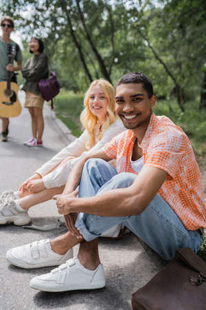 happy african american man smiling at camera while sitting on road near interracial friendsの写真素材