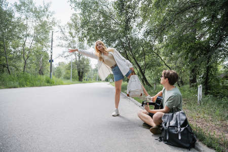 blonde woman stopping car near man sitting on road and playing guitarの写真素材
