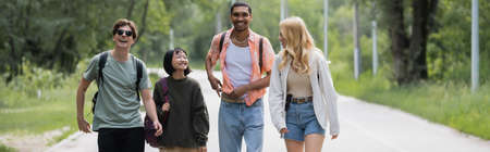 young and cheerful interracial friends walking on road near forest, banner