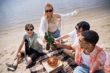 smiling woman pointing with hand while clinking beer bottles with multicultural friendsの写真素材
