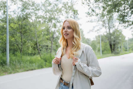 young blonde tourist with backpack smiling on road in countrysideの写真素材