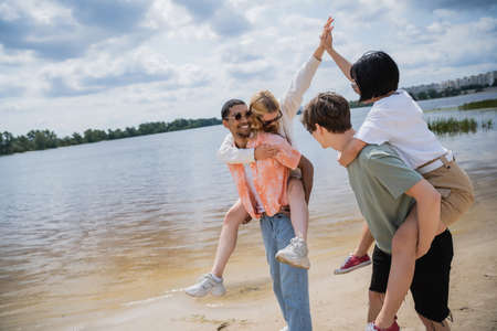 young interracial friends piggybacking women giving high five near river