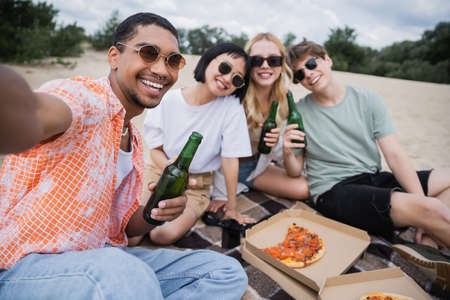 happy multiethnic friends in sunglasses holding beer near tasty pizza on beachの写真素材