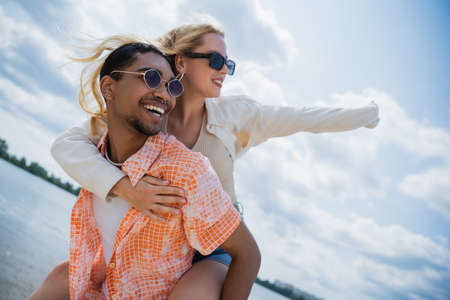happy african american man in sunglasses piggybacking excited girlfriend on summer day