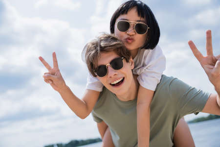 man in sunglasses piggybacking asian girlfriend pouting lips and showing victory gestureの写真素材