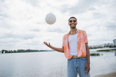 happy african american man in sunglasses looking at camera while playing with ball near riverの写真素材