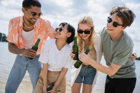 joyful multiethnic friends holding beer bottles on beach