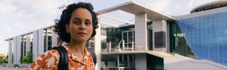 curly young woman with backpack near modern contemporary building in berlin, banner
