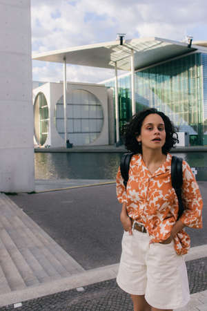 curly young woman standing with hands in pockets near modern contemporary building in berlin