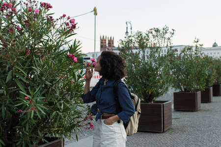 curly young woman with backpack smelling flower and standing with hand in pocket on street in berlin