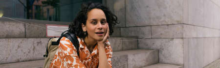 curly young woman in floral shirt sitting on stairs on street in berlin, banner