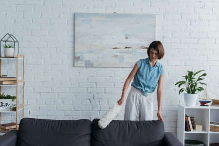 young brunette woman cleaning sofa with dust brush in living room