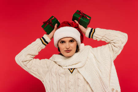 Young woman in santa hat and knitted sweater holding gifts isolated on red