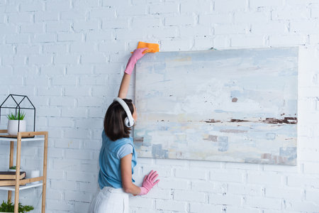woman in rubber gloves and wireless headphones wiping painting with ragの写真素材