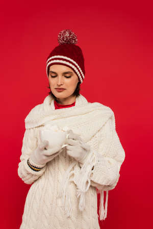 Young woman in warm clothes holding cacao with marshmallows isolated on red