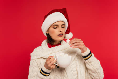 Young woman in santa hat holding cup and marshmallow isolated on red