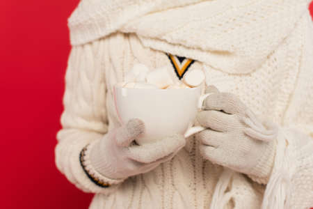 Cropped view of woman in knitted clothes and gloves holding cup with marshmallows isolated on red