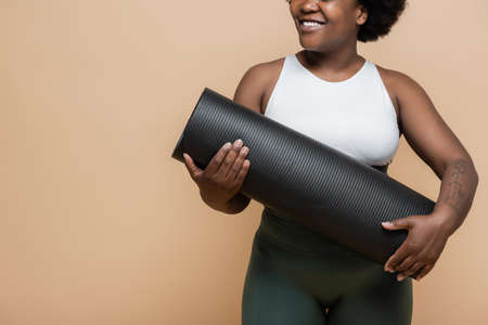 cropped view of joyful african american plus size woman with tattoo holding fitness mat isolated on beigeの写真素材