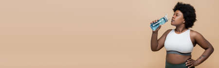 african american plus size woman drinking water and standing with hand on hip isolated on beige, bannerの写真素材