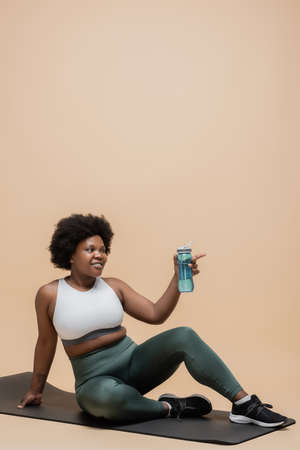 positive african american plus size woman in sportswear sitting on fitness mat and holding sports bottle on beigeの写真素材