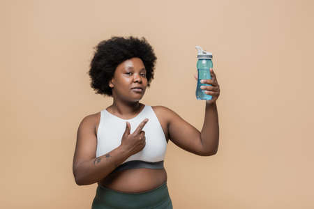 african american plus size woman in crop top holding sports bottle with water and pointing at...