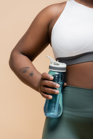 cropped view of tattooed african american plus size woman holding sports bottle isolated on beigeの写真素材