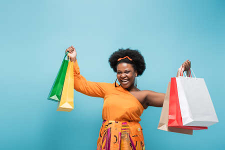 excited african american body positive woman holding shopping bags isolated on blueの写真素材