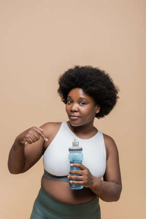 african american plus size woman in crop top pointing at sports bottle with water isolated on beigeの写真素材