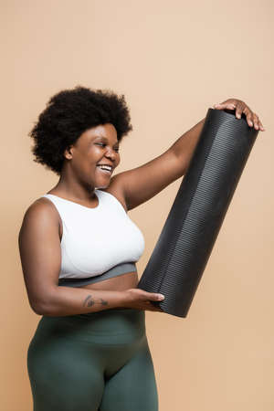 happy african american plus size woman in crop top looking at fitness mat isolated on beigeの写真素材