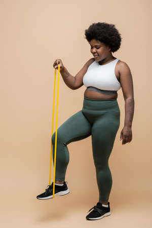 full length of happy african american plus size woman in sportwear exercising with resistance band on beigeの写真素材