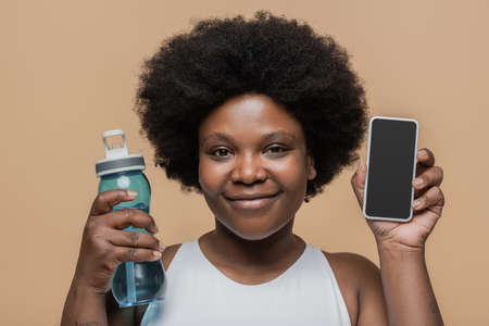 smiling african american plus size woman holding sports bottle and smartphone with blank screen isolated on beigeの写真素材