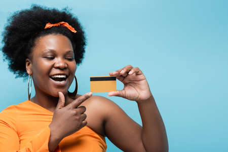 amazed african american body positive woman in hoop earrings pointing at credit card isolated on blueの写真素材