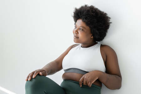 smiling african american plus size woman in sportswear looking away on white