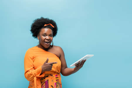 amazed african american body positive woman in hoop earrings pointing at digital tablet isolated on blueの写真素材