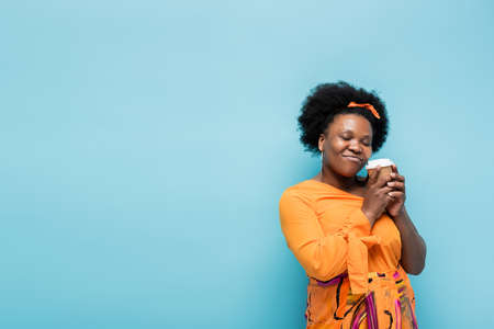 pleased african american body positive woman in hoop earrings holding paper cup isolated on blue