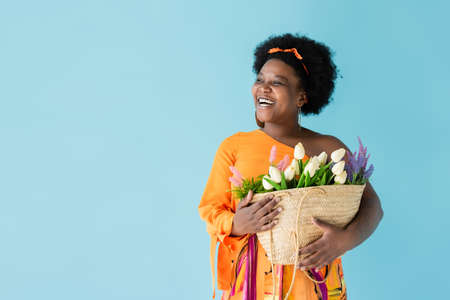 positive african american woman holding straw bag with flowers isolated on blue
