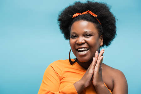 happy african american body positive woman in orange dress and hoop earrings isolated on blueの写真素材