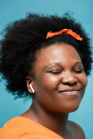 portrait of pleased african american body positive woman in wireless earphone listening music isolated on blueの写真素材
