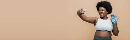happy african american plus size woman holding sports bottle and waving hand during video call isolated on beige, bannerの写真素材