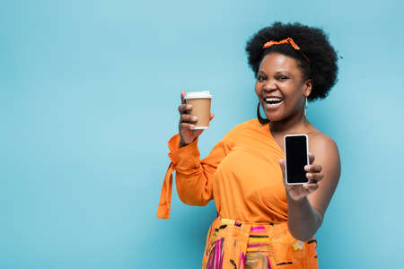 happy african american body positive woman holding paper cup and smartphone with blank screen isolated on blueの写真素材