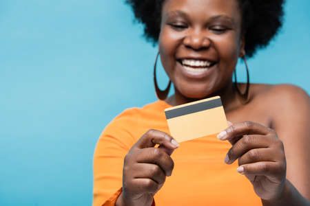 happy african american body positive woman in hoop earrings holding credit card isolated on blue