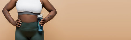 cropped view of tattooed african american plus size woman holding sports bottle with water isolated on beige, bannerの写真素材