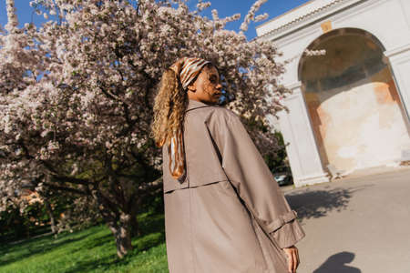 young african american woman in headscarf and stylish trench coat standing near blooming tree in pragueの写真素材