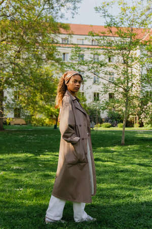 young african american woman in headscarf and stylish trench coat standing with hand in pocket on green grass in parkの写真素材