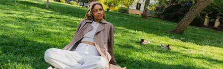 young african american woman in headscarf and stylish trench coat resting on green grass in park,...